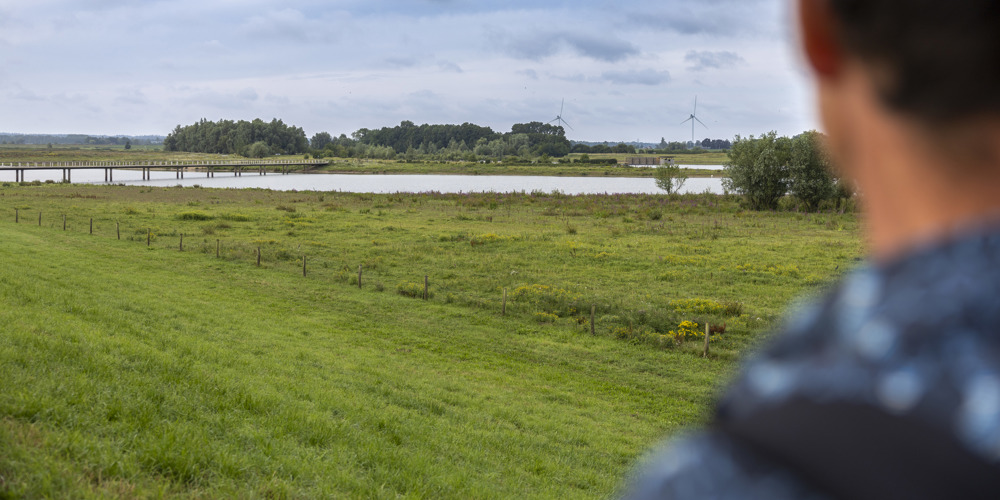 Vaag op de voorgrond staat collega Chris, de achterkant van zijn hoofd is te zien. Op de achtergrond is scherp de Vreugderijkerwaard te zien.