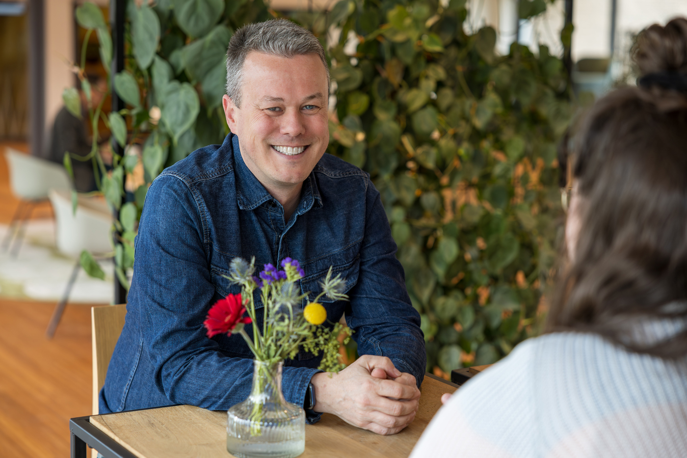 Collega Björn zit aan een tafel en kijkt in de camera. Op de tafel staat een vaasje met wat gekleurde bloemen erin en tegenover Björn zit een vrouwlijke collega.