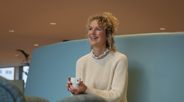 Sandra zit op een blauwe bank en draagt een witte trui en heeft een wit kopje vast.