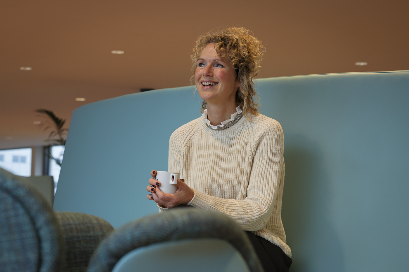 Sandra zit op een blauwe bank en draagt een witte trui en heeft een wit kopje vast.