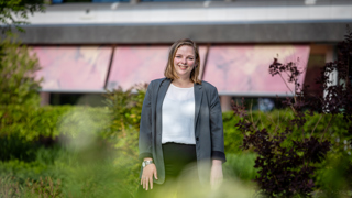 Collega Esther staat in de tuin bij het provinciehuis. Ze stat tussen het groen. Ze draagt een wit shirt en een groene blazer. 
