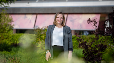 Collega Esther staat in de tuin bij het provinciehuis. Ze stat tussen het groen. Ze draagt een wit shirt en een groene blazer. 