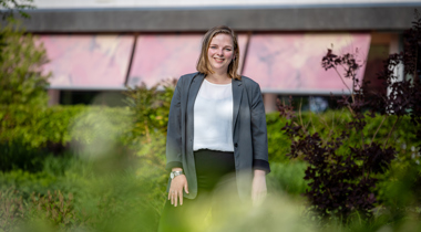 Collega Esther staat in de tuin bij het provinciehuis. Ze stat tussen het groen. Ze draagt een wit shirt en een groene blazer. 