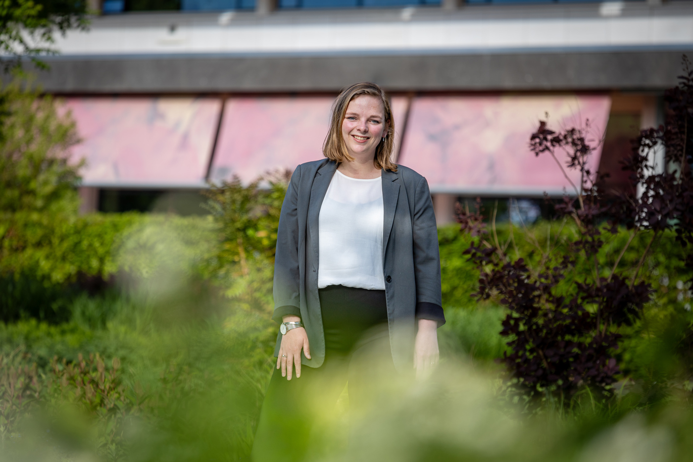 Collega Esther staat in de tuin bij het provinciehuis. Ze stat tussen het groen. Ze draagt een wit shirt en een groene blazer.