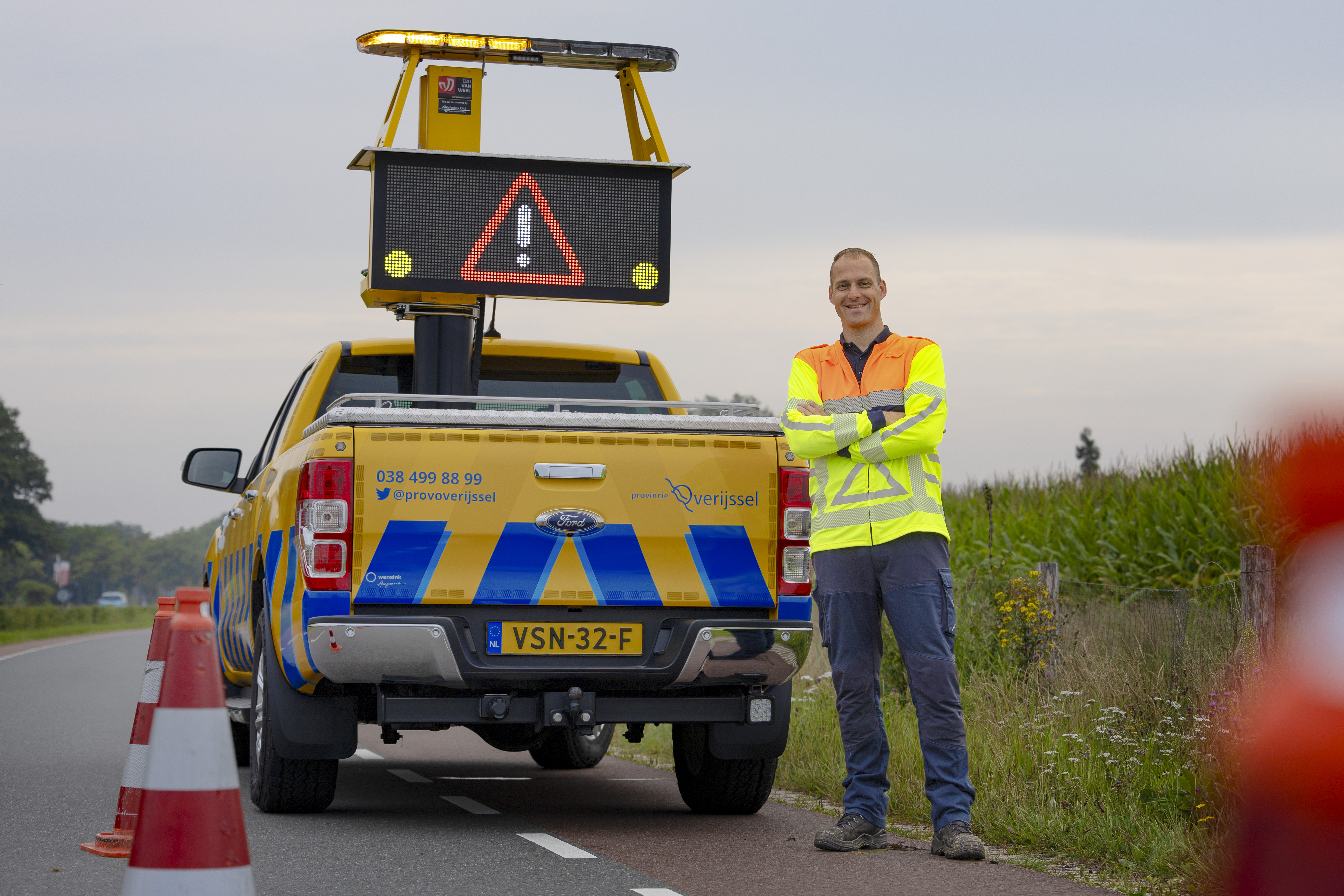 Collega Luuk staat met z'n armen over elkaar voor een geel blauwe provincie auto. Hij staat aan de zijkant van een weg, bij de berm. Hij draagt een oranje geel hesje.