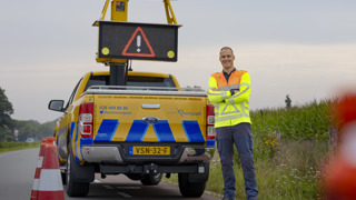 Collega Luuk staat met z'n armen over elkaar voor een geel blauwe provincie auto. Hij staat aan de zijkant van een weg, bij de berm Hij draagt een oranje geel hesje.