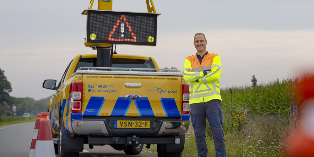 Collega Luuk staat met z'n armen over elkaar voor een geel blauwe provincie auto. Hij staat aan de zijkant van een weg, bij de berm Hij draagt een oranje geel hesje.