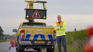 Collega Luuk staat met z'n armen over elkaar voor een geel blauwe provincie auto. Hij staat aan de zijkant van een weg, bij de berm Hij draagt een oranje geel hesje.