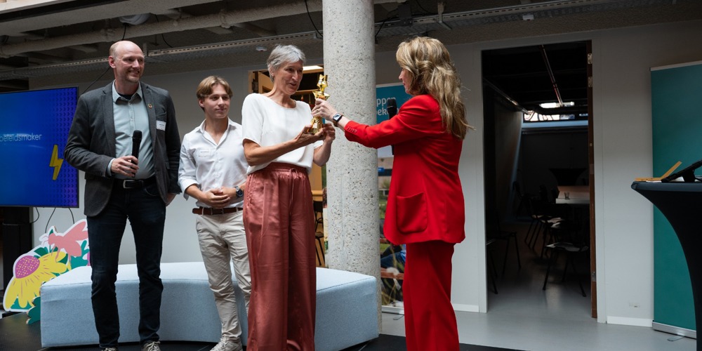 Collega Dicky krijgt een award. Naast haar staan twee mannen en ze krijgt de award van een vrouw met een rood pak aan. Dicky draagt zelf een wit shirt met een roze pantalon.
