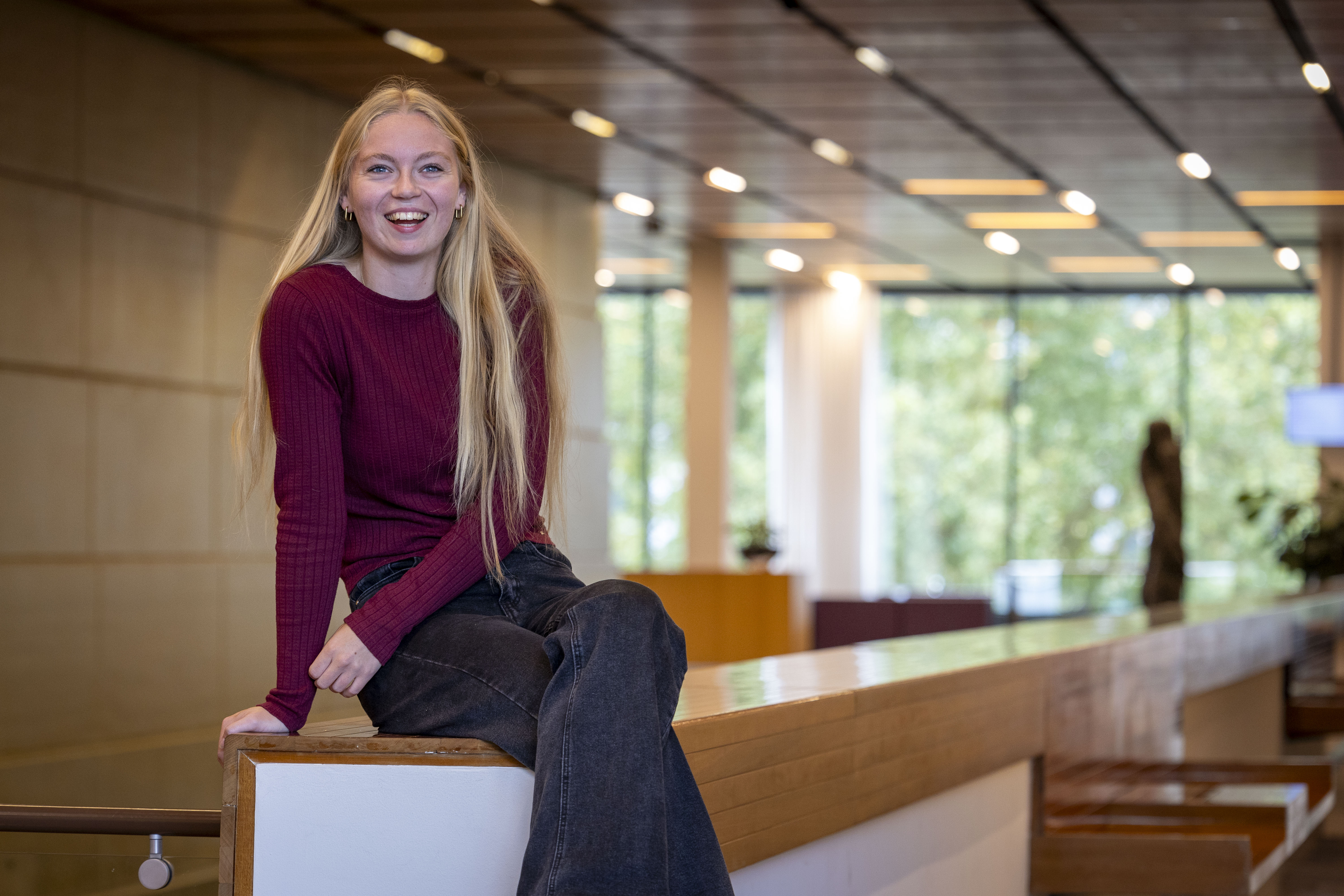 Collega Maaike zit op een houten reling. Ze heeft lang blond haar, draagt een rood tshirt en een zwarte broek.