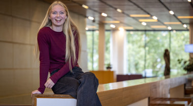 Collega Maaike zit op een houten reling. Ze heeft lang blond haar, draagt een rood tshirt en een zwarte broek.
