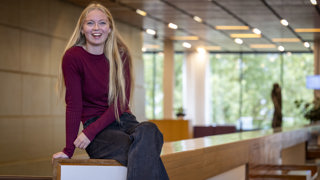 Collega Maaike zit op een houten reling. Ze heeft lang blond haar, draagt een rood tshirt en een zwarte broek.