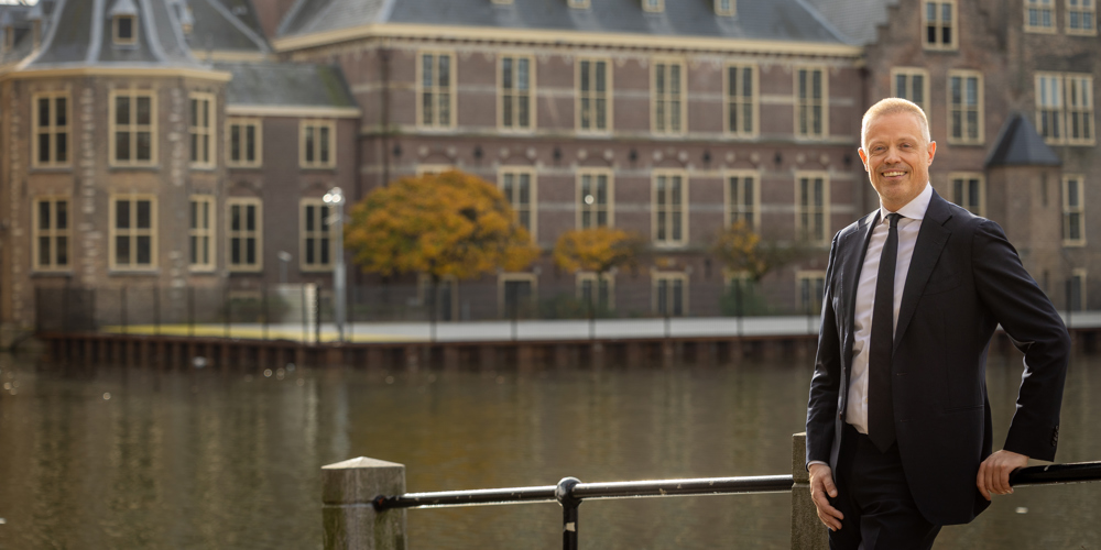 Collega Paul staat tegen een reling aan bij de Hofvijver in Den Haag. Hij draagt een donkerblauw pak. Op de achtergrond is het Binnenhof te zien.