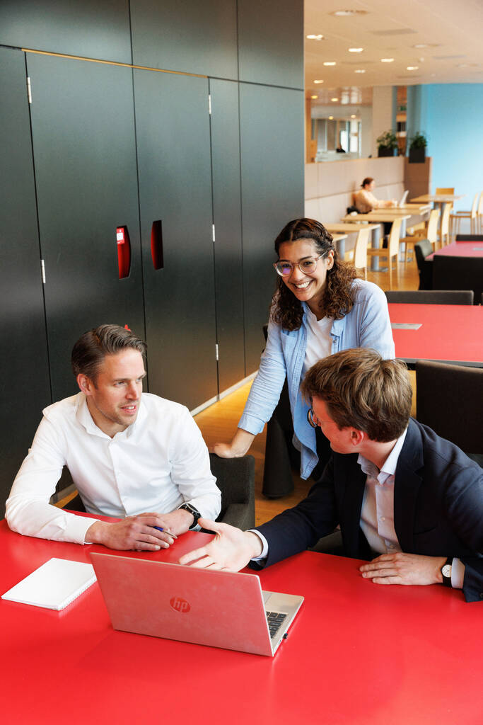 Collega Sharan is in gesprek met twee mannelijke collega's. Zij staat achter de stoelen waar haar collega's op zitten. Ze kijkt de camera in en de collega's kijken naar elkaar. Op de rode tafel waar ze aan zitten staat een laptop open.