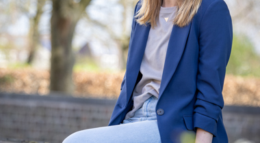 Collega Nathalie zit op een muurtje. Ze draagt een spijkerbroek en heeft beige shirt aan met een blauwe blazer.