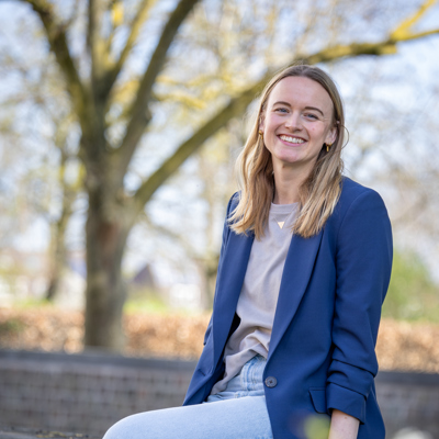 Collega Nathalie zit op een muurtje. Ze draagt een spijkerbroek en heeft beige shirt aan met een blauwe blazer.