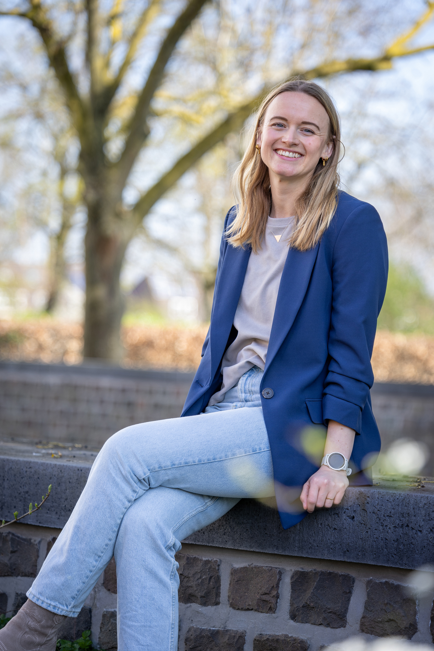 Collega Nathalie zit op een muurtje. Ze draagt een spijkerbroek en heeft beige shirt aan met een blauwe blazer.