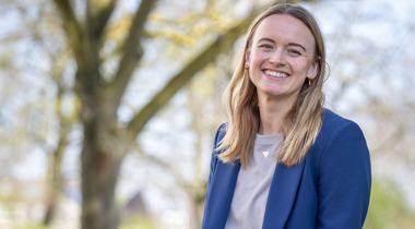 Collega Nathalie zit op een muurtje. Ze draagt een spijkerbroek en heeft beige shirt aan met een blauwe blazer.