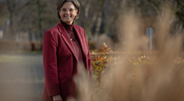 Collega Christa glimlacht naar de camera. Ze staat buiten bij het provinciehuis met bomen en struiken op de achtergrond.