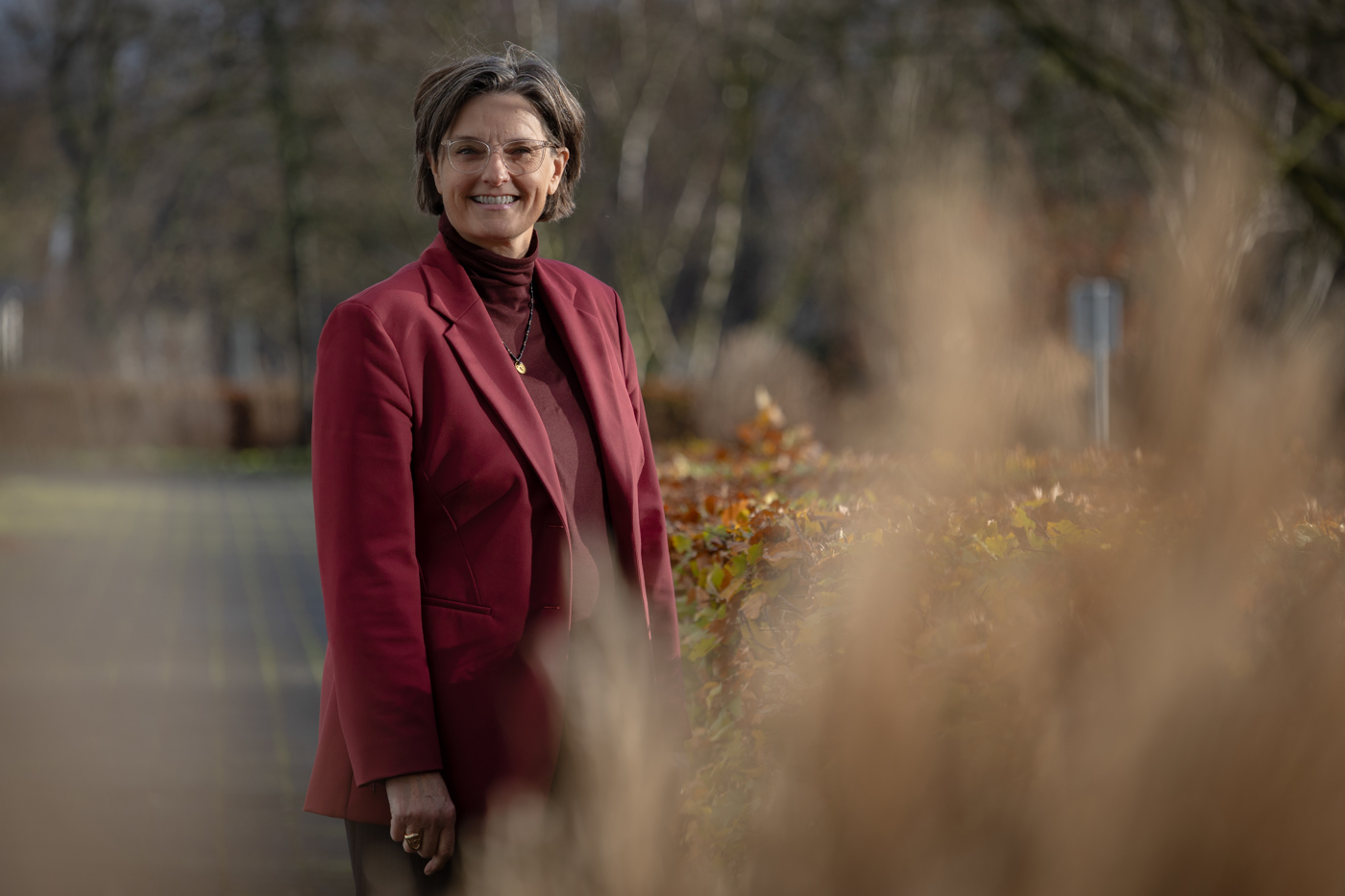 Collega Christa glimlacht naar de camera. Ze staat buiten bij het provinciehuis met bomen en struiken op de achtergrond.