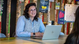 Collega Dorien zit achter een laptop aan een tafel. Ze draagt een blauw gestreepte blouse en een bril.