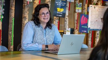 Collega Dorien zit achter een laptop aan een tafel. Ze draagt een blauw gestreepte blouse en een bril.
