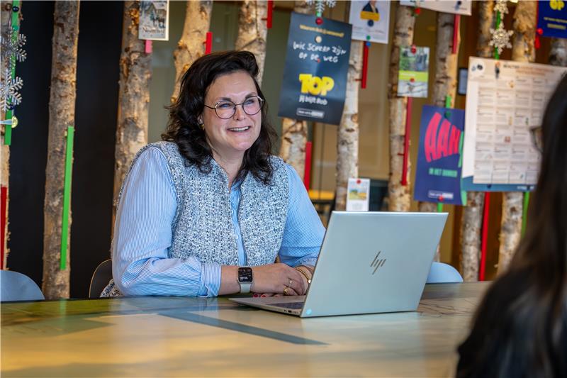Collega Dorien zit achter een laptop aan een tafel. Ze draagt een blauw gestreepte blouse en een bril.