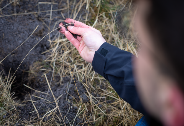 De hand van collega Stijn houdt aarde vast uit een natuurgebied. Verder is er aarde en gras te zien.