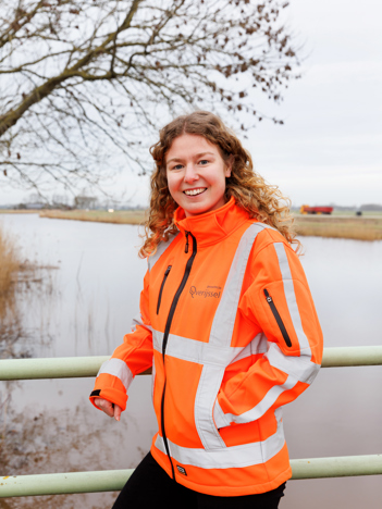 Collega's Maud draagt een oranje hesje en leunt op de reling van een brug en collega Bas staat voor een wit bord met een laptop op een tafel voor hem. Rechts zit in beeld een vrouw.
