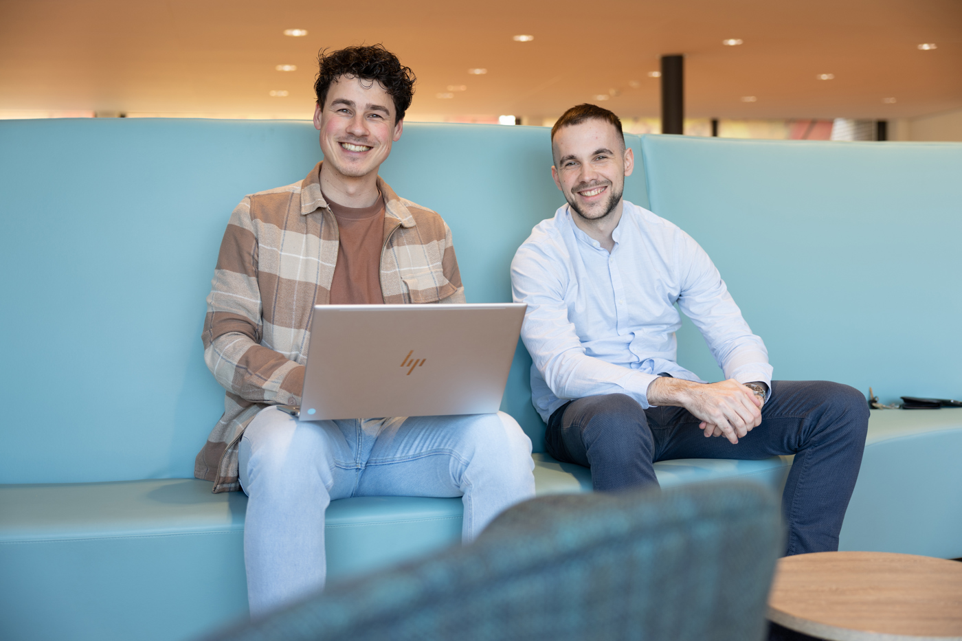 Trainees Tom en Jochem zitten naast elkaar op een blauwe bank. Tom heeft een laptop op zijn schoot.