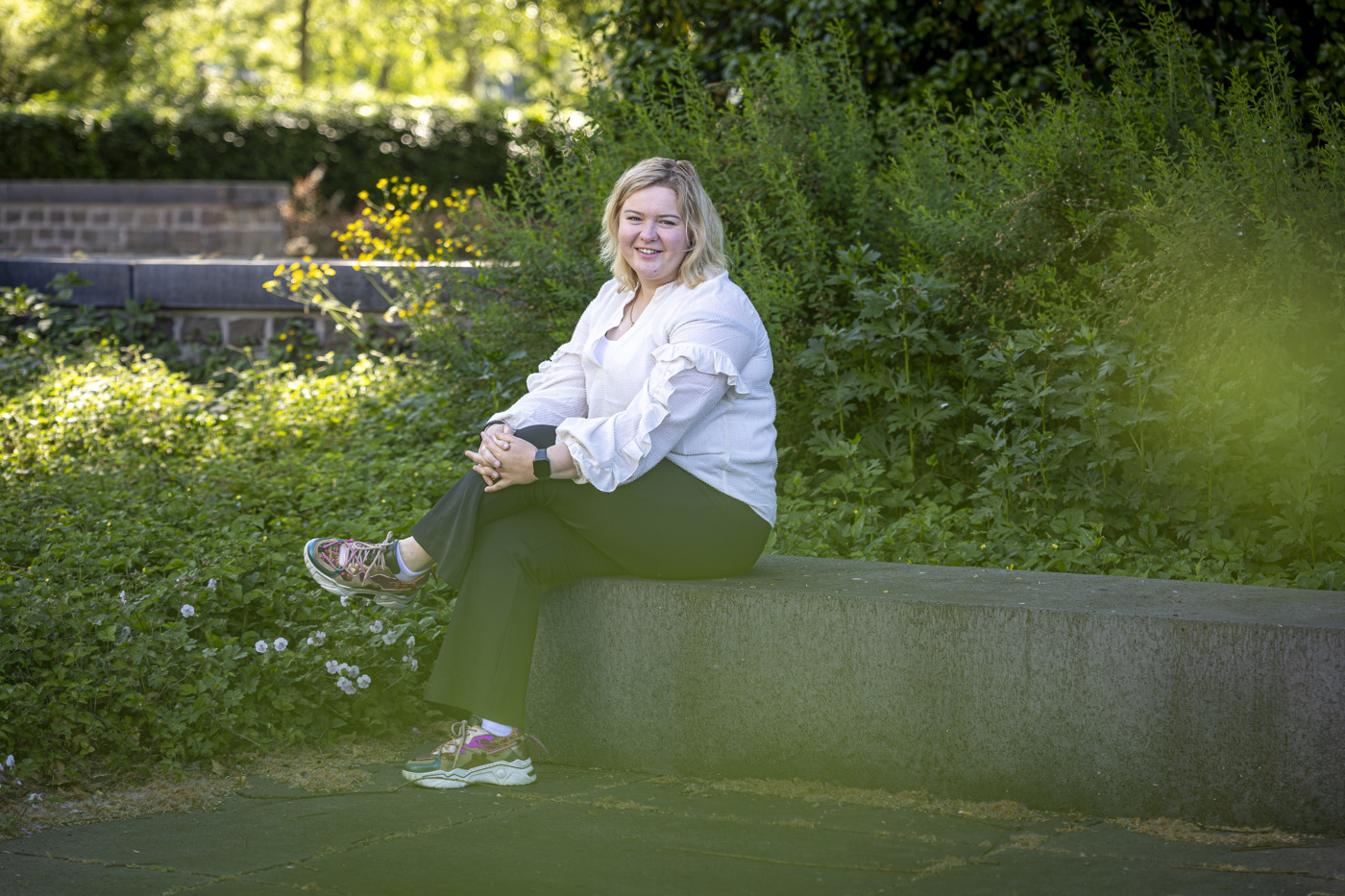 Collega Lizet zit op een stenen bankje in de tuin bij het provinciehuis in Zwolle. Ze draagt een witte blouse en een zwarte broek.