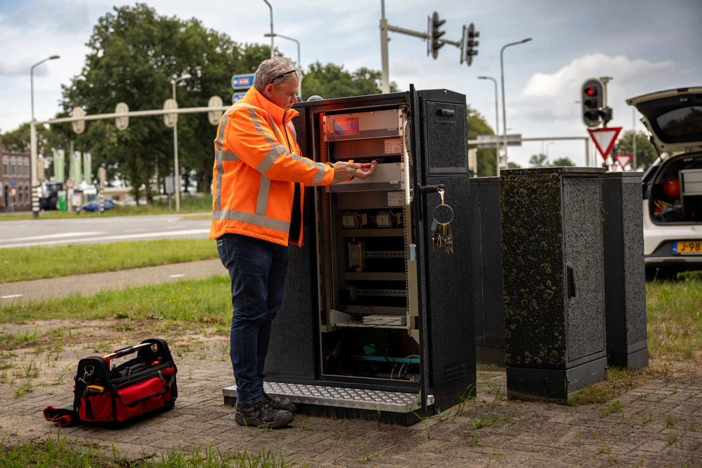Collega Gerdjan werkt aan een verkeersintallatie en draagt een oranje hesje. Op de achtergrond is een auto te zien en een verkeerskruispunt.
