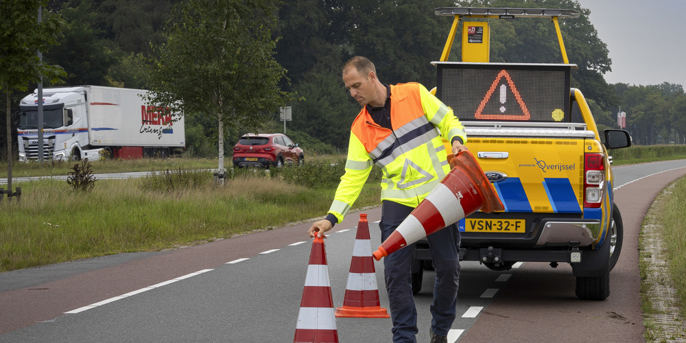 Collega Luuk zet oranje witte pionnen neer op de weg. Hij draagt een oranje geel hesje en achter hem staat een gele provincie auto.
