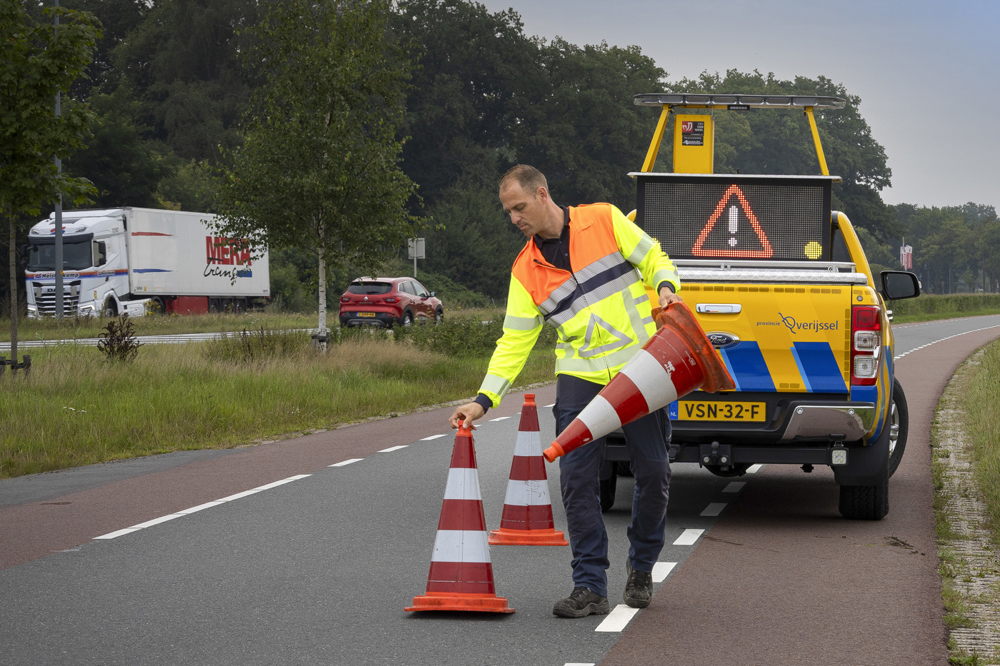 Collega Luuk zet oranje witte pionnen neer op de weg. Hij draagt een oranje geel hesje en achter hem staat een gele provincie auto.