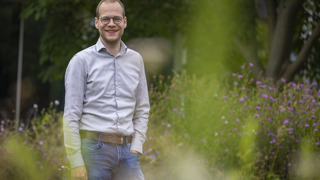 Collega Mart kijkt naar de camera en staat tussen het groen en bloemen. Hij draagt een licht grijs overhemd en een spijkerbroek.