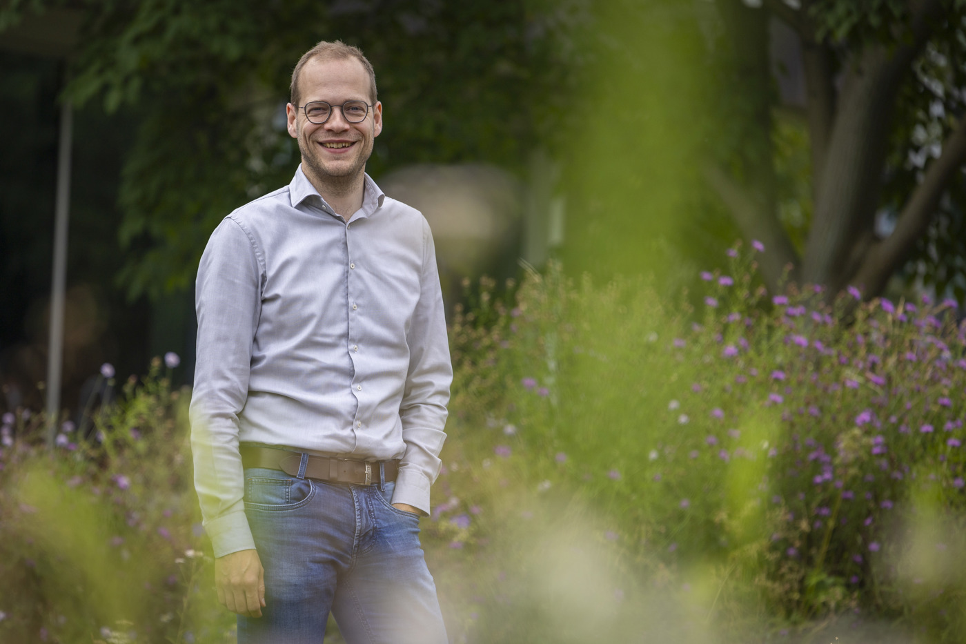 Collega Mart kijkt naar de camera en staat tussen het groen en bloemen. Hij draagt een licht grijs overhemd en een spijkerbroek.