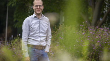 Collega Mart kijkt naar de camera en staat tussen het groen en bloemen. Hij draagt een licht grijs overhemd en een spijkerbroek.
