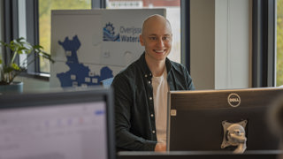 Collega Jeroen zit achter een computerbeeldscherm. Hij zit dichtbij ramen en op de achtergrond is een bord van de Overijsselse Wateraanpak te zien waar de vorm van de provincie Overijssel te zien is.