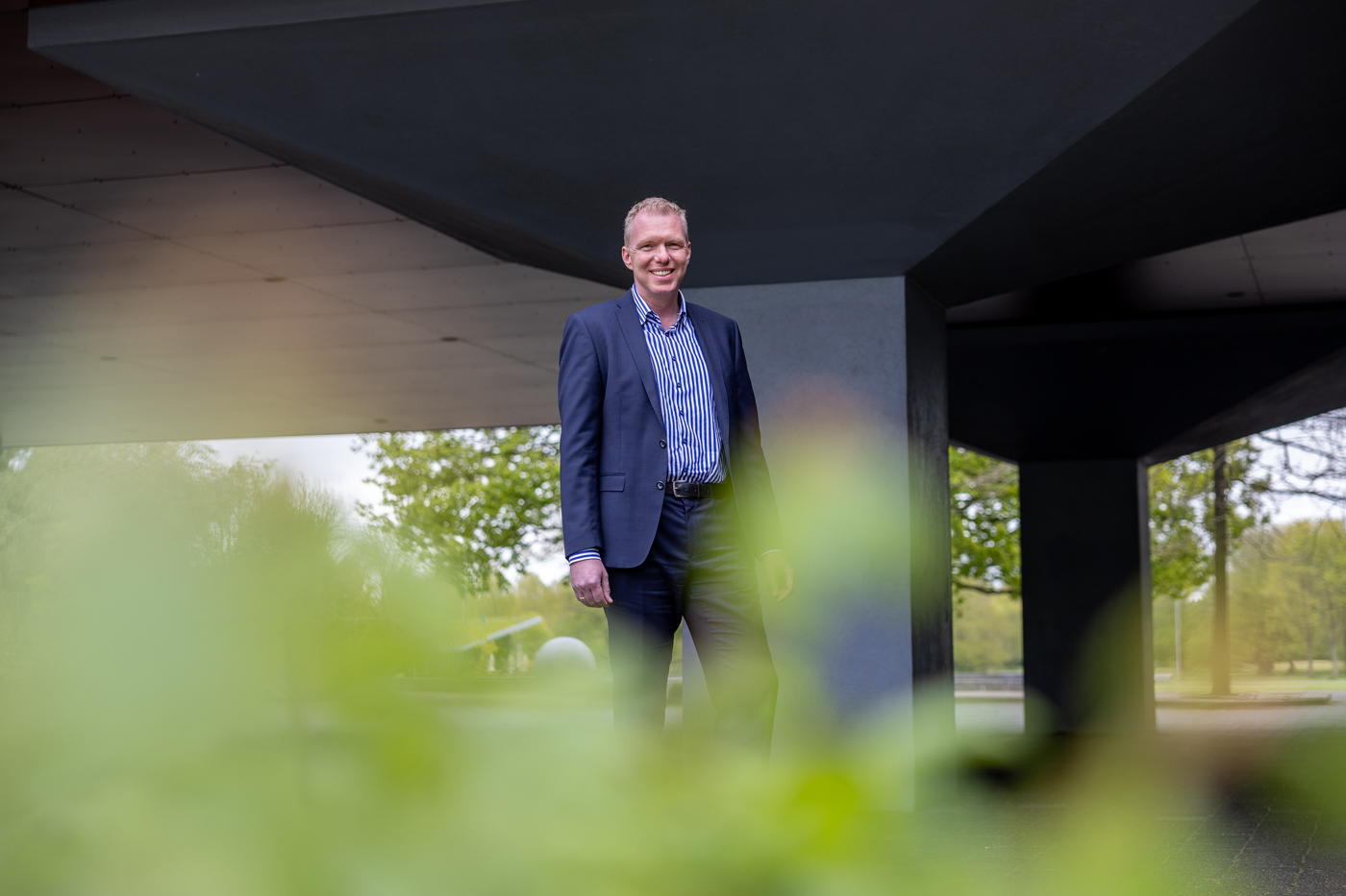 Een portret foto van collega Henri die buiten voor het provinciehuis staat. Op de voorgrond van de foto zijn wat groene bladeren te zien.