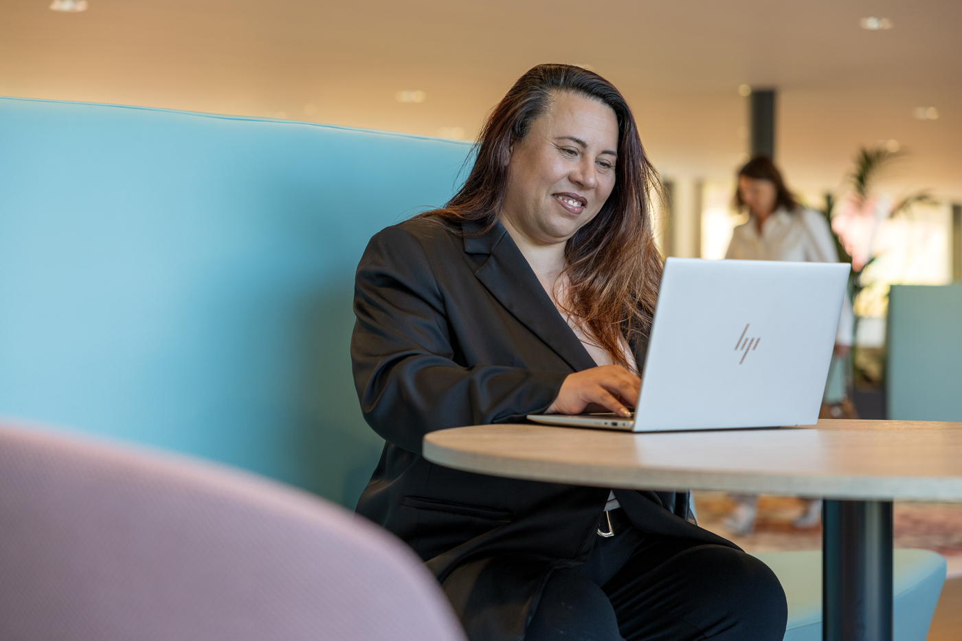 Collega Melanie zit op een blauwe bank in het provinciehuis te werken achter haar laptop.