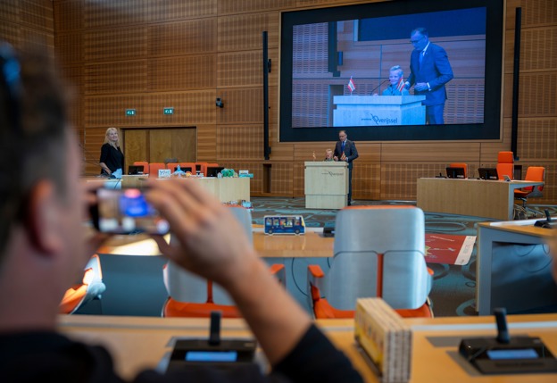 De stagenzaal van Overijssel is te zien. Op het scherm staat een man met een jongen achter de microffon. Verschillende stoelen van de statenzaal zijn te zien. Op de voorgrond staat een man te filmen met zijn telefoon.