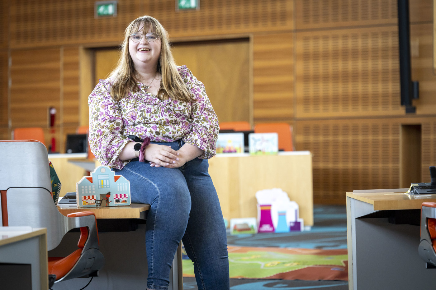 Collega Vianna zit in de statenzaal. Ze leunt op een tafel en achter haart zijn houten attributen te zien die gebruikt worden tijdens het provinciespel.