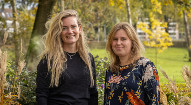 Collega's Lisa en Janine staan buiten voor een groene omgeving. Lisa heeft blond haar en draagt een zwart shirt. Janine heeft blond haar en draagt een shirt met bloemen.