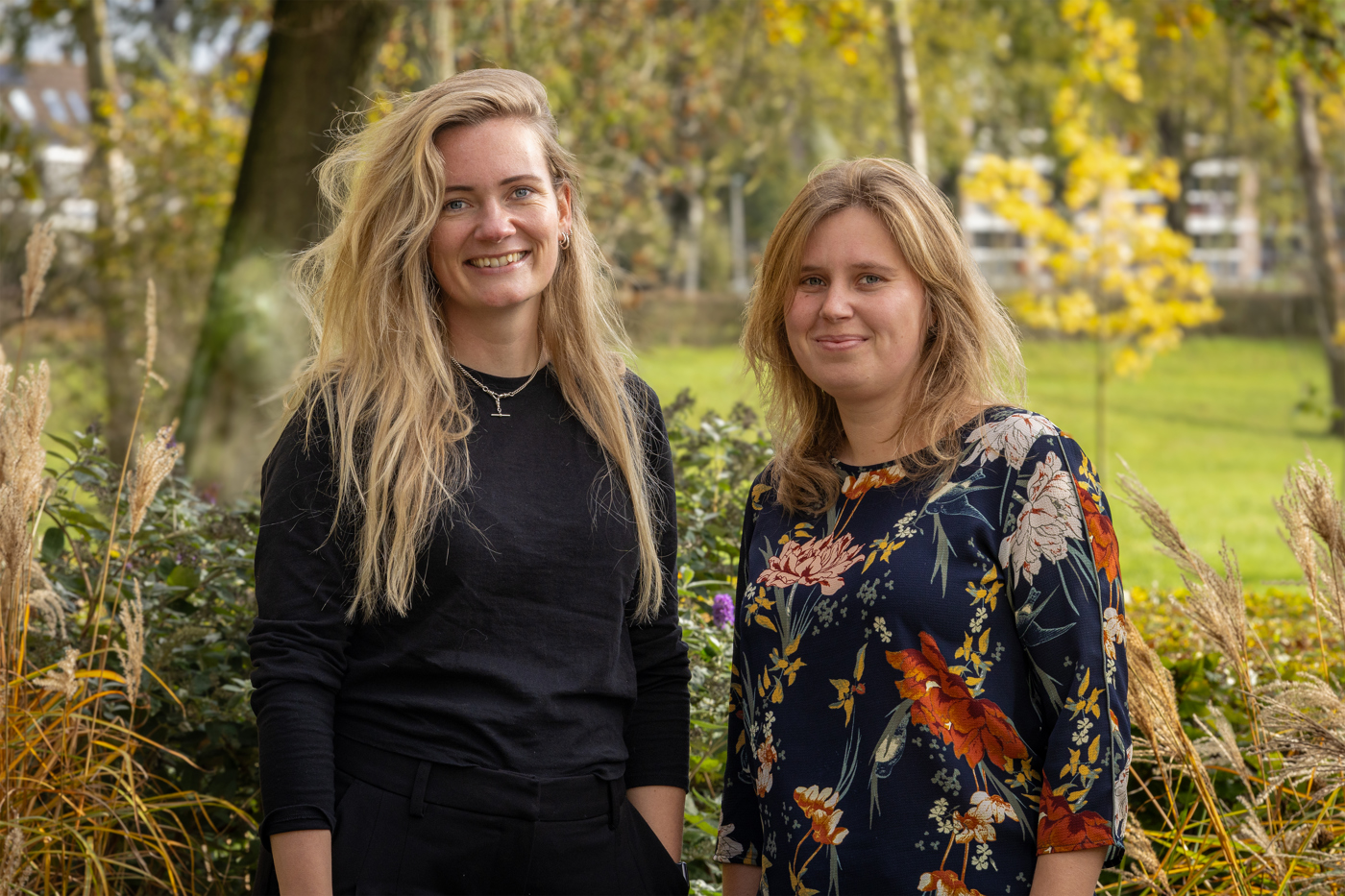 Collega's Lisa en Janine staan buiten voor een groene omgeving. Lisa heeft blond haar en draagt een zwart shirt. Janine heeft blond haar en draagt een shirt met bloemen.