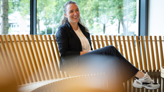 Collega Joanne zit op een bruine ronde houten bank. ze draagt een donkerblauw pak met een witte blouse en sneakers.