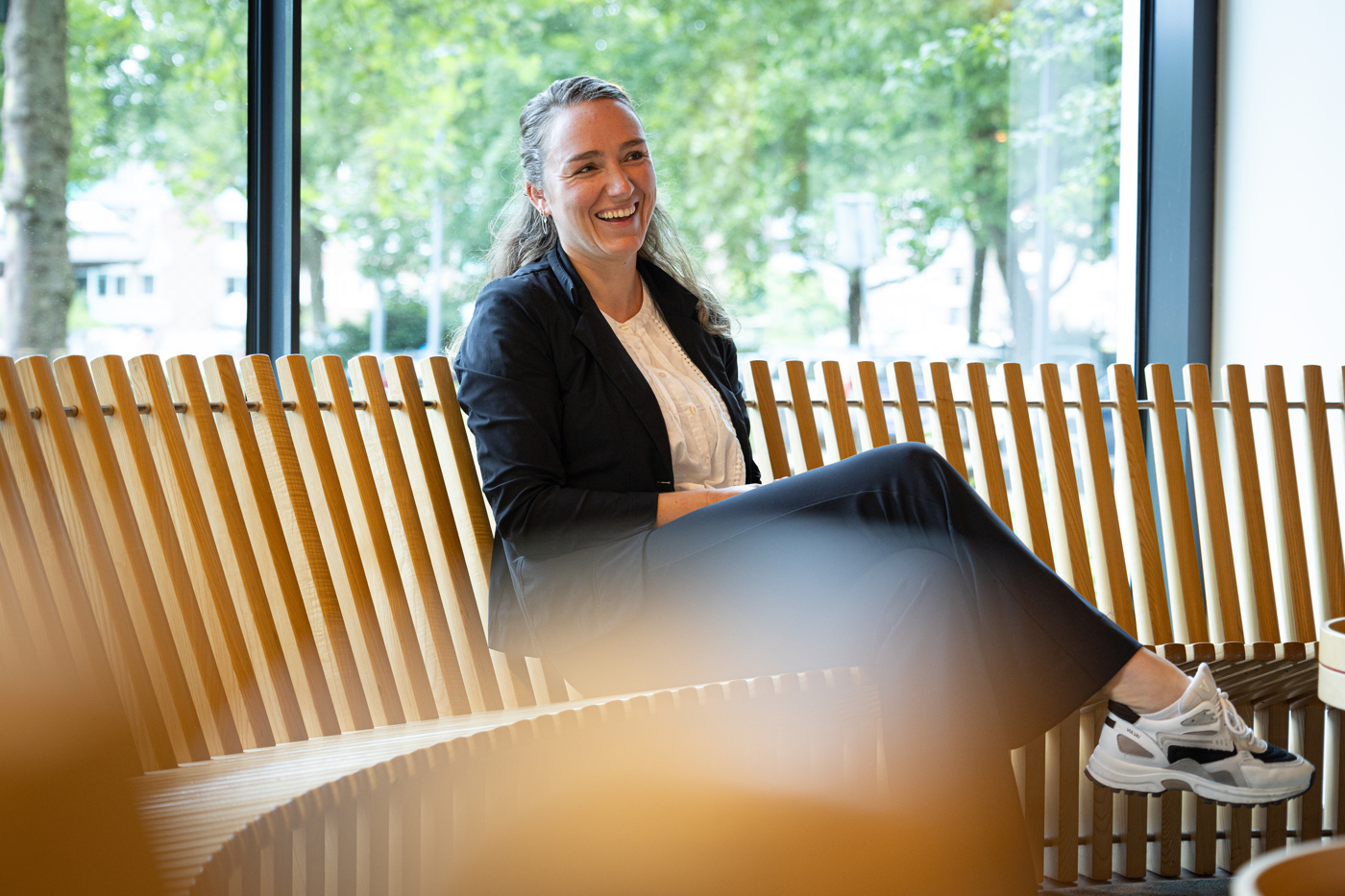 Collega Joanne zit op een bruine ronde houten bank. ze draagt een donkerblauw pak met een witte blouse en sneakers.
