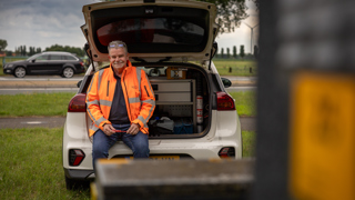 Collega Gerdjan draagt een oranje hesje en zit in de achterbank van een auto.