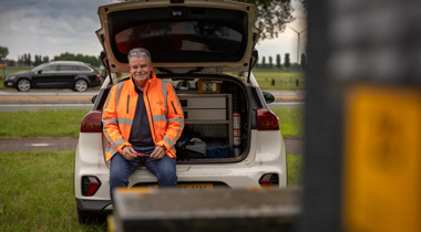 Collega Gerdjan draagt een oranje hesje en zit in de achterbank van een auto.