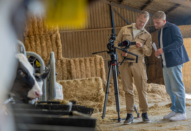 Op de eerste afbeelding staat Koen buiten het provinciehuis bij een boom, hij draagt een grijze trui. Op de tweede afbeelding staat hij met een cameraman bij een statief in een stal. Op de voorgrond is een koe te zien.