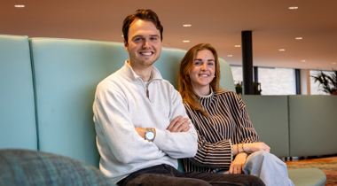 Collega's Nick en Laura zitten naast elkaar op een blauwe bank. Collega Nick draagt een gruize trui en een zwarte broek. Collega Laura draagt een zwart wit gestreepte trui en een spijkerbroek.